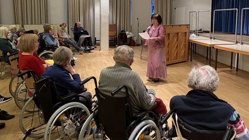 Mehrere alte Menschen im Rollstuhl hören einer Sängerin zu, die ein rosa Kleid trägt.